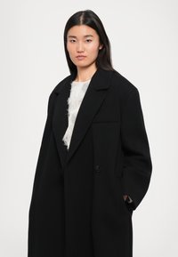 Černý oversized kabát s klopovým límcem, zapínáním na jeden knoflík a bočními kapsami, vrstvený přes texturovaný bílý top.