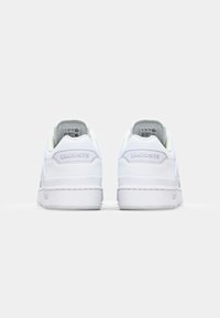 Białe skórzane trampki o gładkiej fakturze, z wyściełanym kołnierzem i logo Lacoste. Płaska gumowa podeszwa o minimalistycznym designie. Widok z tyłu.