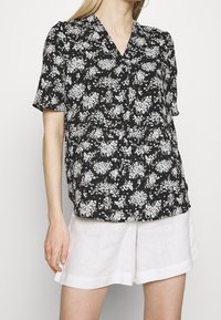 Blouse noire à manches courtes avec un motif floral en bleu clair et beige, design en v, coupe décontractée, associée à un short en lin blanc.