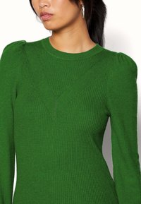 Zielony sweter z prążkowanego materiału, o dopasowanym kroju, z okrągłym dekoltem i ukośnymi szwami na klatce piersiowej oraz ramionach.