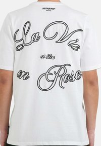 T-shirt bianco in cotone con testo ricamato nero sul retro: "La Vie en Rose" con elementi calligrafici e decorativi. Design semplice.