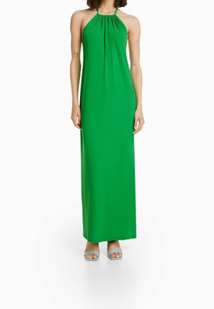 Groene maxijurk met halternek, een geplooide halslijn, een gladde textuur en een ontwerp tot op de enkels; gedragen met lichtblauwe sandalen met bandjes.