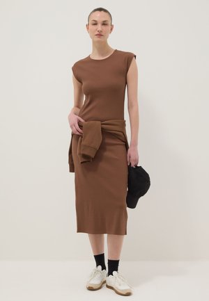 Mujer con vestido marrón sin mangas y sudadera marrón anudada, sosteniendo una gorra negra y zapatillas blancas con calcetines negros.