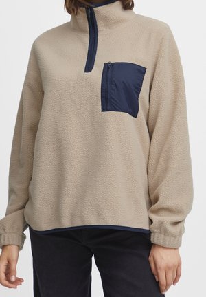 Beige fleece pullover met marineblauwe rits tot de helft en een marineblauwe ritszak op de borst, gedragen met zwarte broek.
