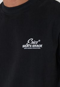 Camiseta negra de cuello redondo con texto blanco "Erics Skate Shack Santa Monica Boulevard" en el pecho, usada por una persona.