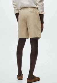 Pantaloni corti beige in cotone con vita elasticizzata, tasca posteriore e vestibilità rilassata, abbinati a scarpe marroni senza lacci su uno sfondo neutro.