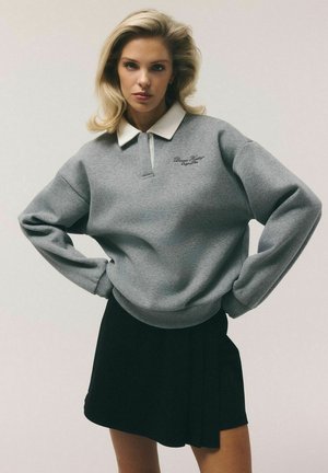 Młoda kobieta o blond włosach nosi szarą bluzę z białym kołnierzem i czarną spódnicę, stojąc z rękami na biodrach na jednolitym tle.