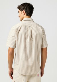 Camicia a maniche corte beige con strisce verticali marroni chiare, dotata di una piega sul retro, colletto a bottone e un piccolo etichetta logo sul lato.