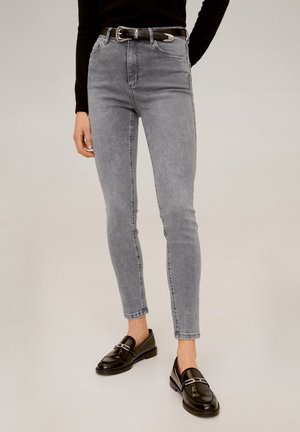 Persona que lleva vaqueros ajustados grises, cinturón negro con hebilla plateada, camiseta negra de manga larga y mocasines de cuero negros con adornos plateados.