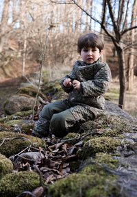 Pehmendusega kaetud rohelise maskivaate jope ja püksid lillemustriga, istub samblasel kivil okste ja lehtede keskel metsas.