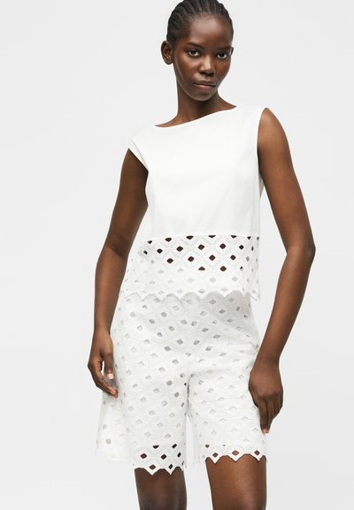 Γυναίκα που φοράει ένα αμάνικο λευκό τοπ με γεωμετρική δαντέλα στο τελείωμα και αντίστοιχα σορτς μέσης δαντέλας μέχρι το γόνατο, όρθια μπροστά σε μονόχρωμο φόντο.