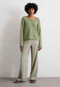 Helles grünes Strickoberteil mit lässiger Passform und weit geschnittenen Ärmeln, kombiniert mit glatten, helloliven, weiten Hosen und flachen schwarzen Sandalen.