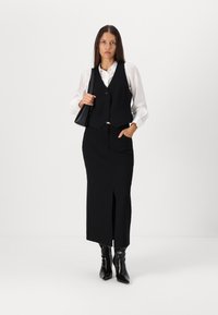 Black vest with a single button, layered over a white shirt. Black fitted skirt with a slit and black ankle boots. Minimalist accessory included.