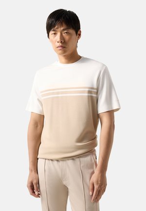 Mann trägt ein beige-weißes kurzärmeliges Hemd mit horizontalen Streifen und hellbeiger Hose, steht vor einem einfarbigen Hintergrund.