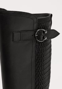 Botte noire avec un panneau tissé texturé, une tige en cuir lisse et une sangle avec une boucle en métal circulaire pour le détail.