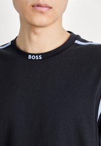 Fekete rövid ujjú póló, kerek nyakkivágással, világoskék részletekkel a vállakon, és a "BOSS" szó világoskék színben a nyakkivágásnál.