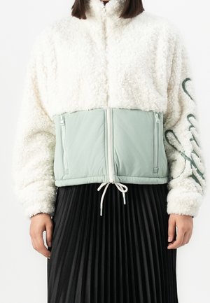 Personne portant une veste en polaire sherpa blanche avec des poches zippées vert clair et une jupe plissée noire.