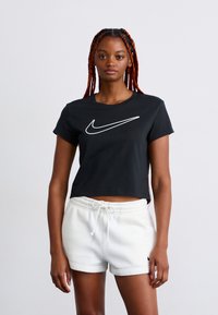 Jovem mulher com tranças vermelhas, vestindo uma t-shirt preta corta da Nike e calções brancos com cordão, contra um fundo simples.