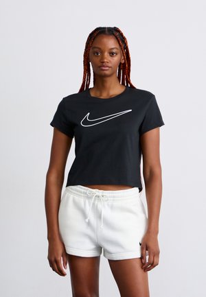 Ung kvinne med røde fletter som har på seg en svart cropped Nike t-skjorte og hvite shorts med snøring foran en ensfarget bakgrunn.