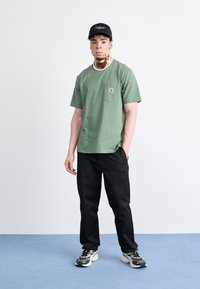 Camiseta de algodón verde con bolsillo y logo, combinada con pantalones negros y zapatillas con un patrón de camuflaje y detalles en blanco. Se incluye una gorra negra.
