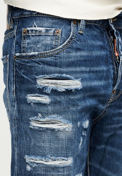 Gros plan sur un jean en denim bleu usé avec plusieurs déchirures horizontales et des bords effilochés au niveau des cuisses et des poches.