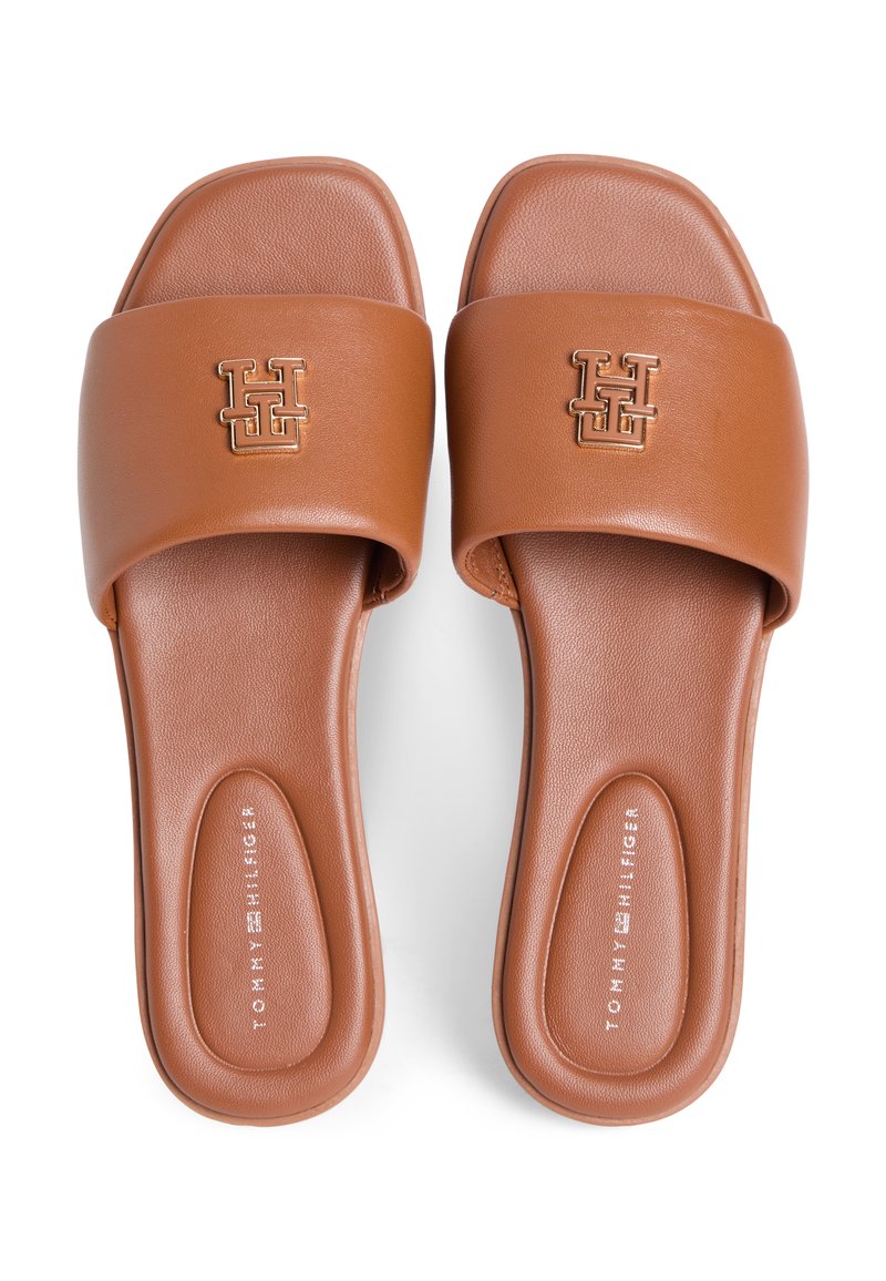 Sandalias tipo slide sintéticas marrones con una correa ancha que presenta un logo dorado, plantilla texturizada y superficie lisa. No se ven detalles de la suela.