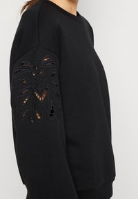 Felpa nera con maniche lunghe caratterizzata da intricati motivi a taglio sulla manica sinistra, che creano una texture stratificata e un dettaglio di design.