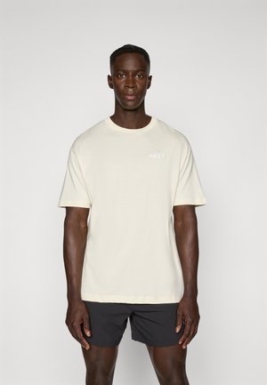 Cremefarvet T-shirt med korte ærmer, rund halsudskæring og et lille broderet logo. Bæret med mørke shorts. Glat bomuldsstruktur.