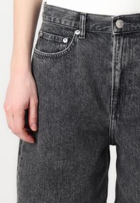 Jean en denim noir à taille haute, design à cinq poches et quincaillerie en ton argenté. La texture semble légèrement délavée avec des détails de lavage subtils.