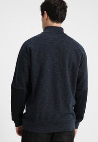 Uomo che indossa un pullover a maniche lunghe grigio scuro e nero visto da dietro su uno sfondo bianco uniforme.