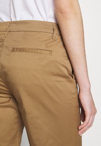 Beżowe spodnie o gładkiej fakturze, z pojedynczą tylną kieszenią i detalem z logo na pasku, prezentowane na dłoni z boku.