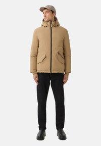 Beige Steppjacke mit Kapuze, Reißverschluss, zwei Fronttaschen und gerippten Bündchen. Kombiniert mit dunklen Jeans und schwarzen Slippers.