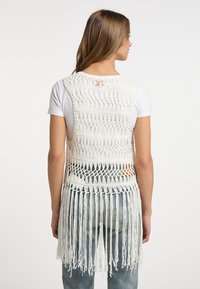 Gilet bianco all'uncinetto con frange lunghe, motivo a trama aperta e design senza maniche, indossato sopra una maglietta bianca e abbinato a pantaloni di denim.