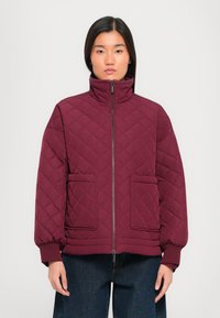 Burgundy quilted jacket with a high collar, front zip, and two large patch pockets; features ribbed cuffs and a textured finish.