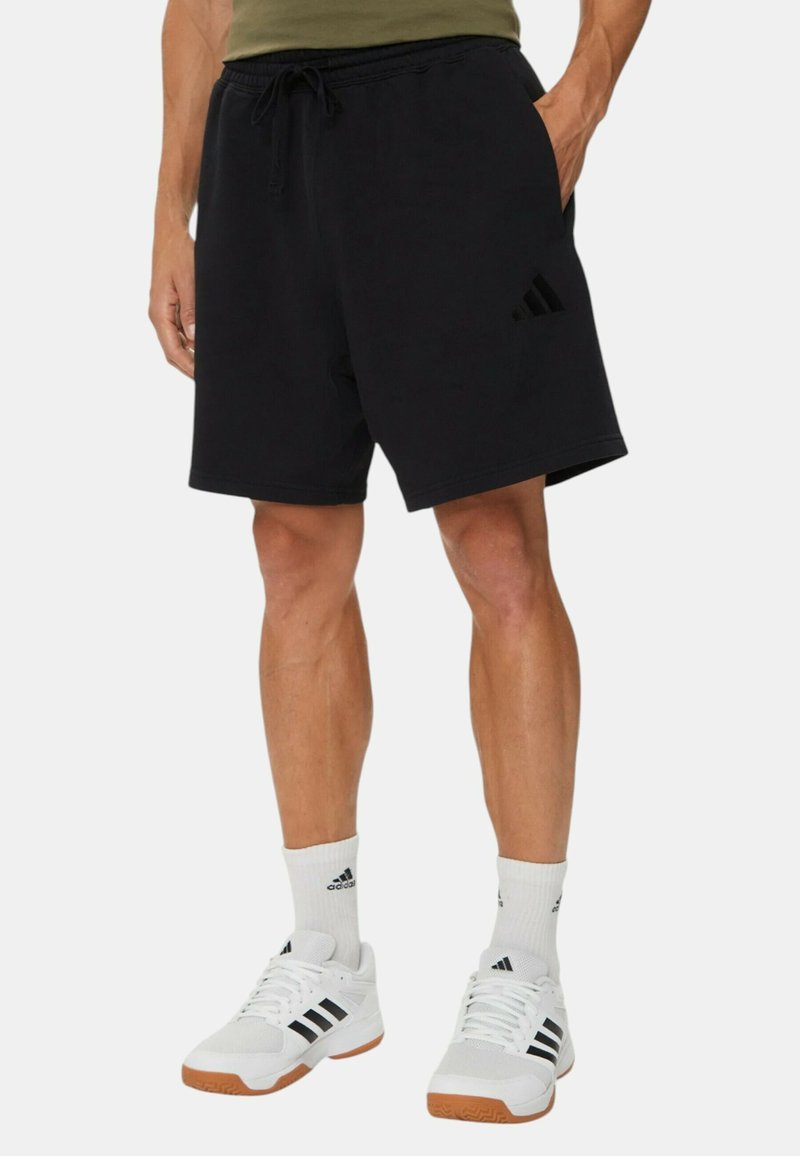 Pantalones cortos deportivos negros con una banda elástica en la cintura y cordón, que presentan un sutil detalle del logo de Adidas en el lateral, combinados con zapatos blancos.