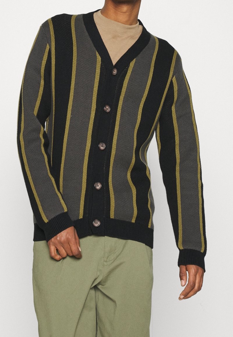 Homme portant un cardigan rayé vertical noir et gris avec des lignes jaunes et un pantalon vert sur fond blanc.