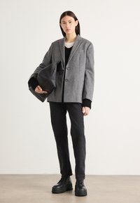 Blazer en laine gris avec deux boutons, porté sur un top en tricot noir, associé à un pantalon noir et des bottes noires épaisses. Design simple et moderne.
