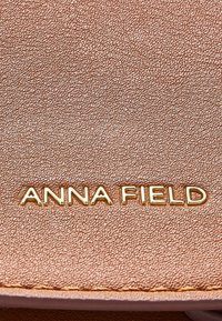 Material texturizado de oro rosa con el nombre de la marca "ANNA FIELD" en relieve en letras doradas. Detalle en primer plano de la superficie.