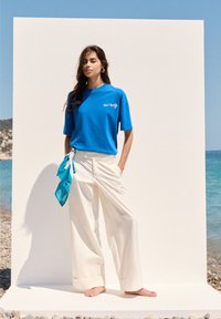 Blå bomulds t-shirt med hvidt logo, parret med hvide brede bukser. Et lyseblåt tørklæde er bundet om livet. Baggrunden viser en strandscene.