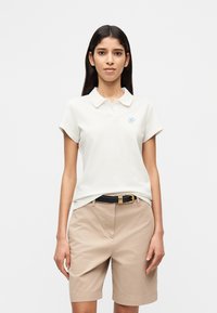 Λευκό πόλο πουκάμισο με διπλωμένο γιακά και κοντά μανίκια, με λογότυπο σε ανοιχτό μπλε χρώμα. Συνδυάζεται με μπεζ σορτς και μαύρη ζώνη.