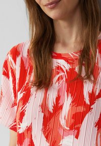 Rood en wit geplooid top met een bloemenpatroon. Het ontwerp heeft korte mouwen en een ronde halslijn. Glad textuur materiaal.