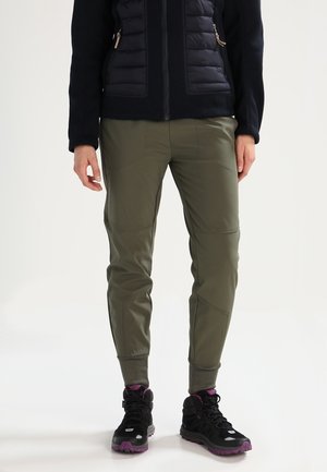 Personne portant un pantalon fuselé vert olive, une veste noire avec fermeture éclair et des chaussures de randonnée noires avec semelles et lacets violets, debout sur un fond blanc.