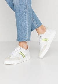 Baskets en cuir blanc avec un bout en suède gris, ornées d'accents à rayures vertes et noires, posées sur une semelle en caoutchouc blanche texturée.