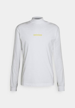 Valkoinen pitkähihaisen mock neck -paita, jossa pieni keltainen vaakasuora raita ja hillitty logosuunnittelu keskellä edessä.