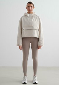 Veste courte beige avec capuche, poche avant et fermeture à pression, portée sur des leggings marron clair et des baskets blanches. Matière lisse, design minimaliste.