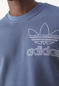 Modrá bavlněná mikina s ribovaným límcem, vyšitou bílou Adidas logem a designem tří pruhů na logu. Hladká textura.