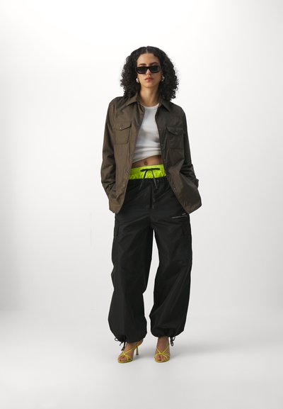 Olivengroene overhemd met knopen, witte cropped top, zwarte cargobroek, neongroene trekkoordtailleband en gele sandalen met bandjes, gestyled met een zonnebril.