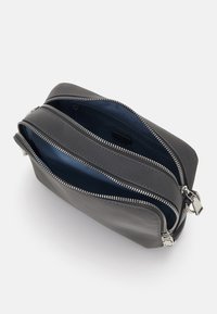 Šedá kožená crossbody taška se dvěma zipovými přihrádkami, stříbrnými kovovými prvky a modrou podšívkou. Hladký povrch a obdélníkový tvar.