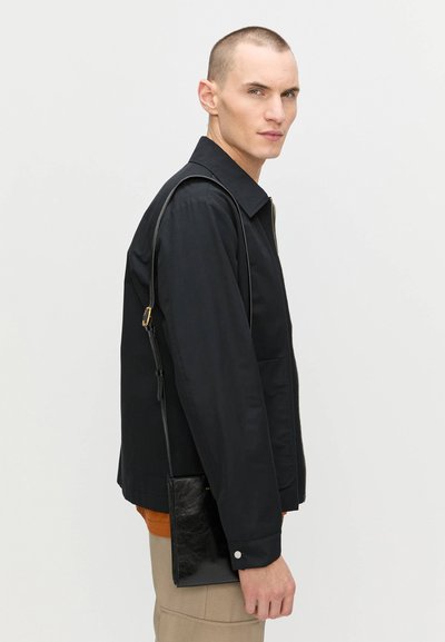 Sort zip-up jakke med spids krave, to forlommer og en moderne pasform; ledsaget af en lille sort taske med skulderrem.