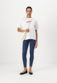 Valkoinen puuvillapaita, jossa on punainen ja sininen graafinen kuvio, yhdistettynä sinisiin slim fit -farkkuihin ja mustiin ballerinan kenkiin. Asusteina on vaalean beige olkalaukku.